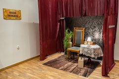 Dawne atelier fotografa w postaci kącika przygotowanego do fotografii portretowej. Jest krzesło, duży wazon z zielonym wiszącym kwiatem, stolik z lampką i akcesoriami, tło.  Z boku na ścianie stylizowany szyld z napisem „Atelier fotografa”