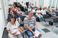 Ponad czterdzieści osób na widowni. Większość siedzi, niektóre osoby w oddali stoją.