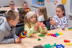 Kilkoro dzieci siedzi przy wspólnych stołach wykonuje ozdoby z bibuły