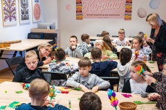 Kilkanaścioro dzieci siedzi przy wspólnych stołach wykonuje palmy wielkanocne z bibuły. Pomagają im kobiety