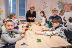 Kilkoro dzieci siedzi przy stole wykonuje lub trzyma palmy wielkanocne z bibuły. Z drugiej strony stołu stoi kobieta, która im  pomaga.
