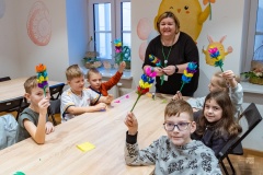 Kilkoro dzieci siedzi przy stole trzyma palmy wielkanocne z bibuły. Z drugiej strony stołu stoi kobieta, która pomagała im w tworzeniu..