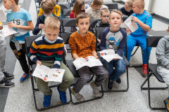 Grupa dzieci  siedzi na krzesłach trzymając w dłoniach foldery oraz kartki z papieru czerpanego
