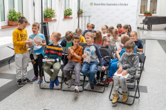 Grupa dzieci  siedzi na krzesłach trzymając w dłoniach foldery oraz kartki z papieru czerpanego