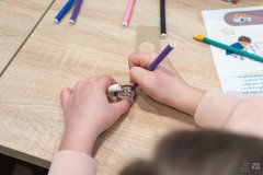 Dłonie dziecka rysujące flamastrem wewnątrz kapsla od butelki serce. Obok leżą inne flamastry oraz karta edukacyjna
