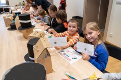 Przy długim stole siedzą dzieci i rozwiązują karty edukacyjne. Na stole  dużo tekturowych kuferków oraz czarnych, papierowych cylindrów i kapeluszy. Dwoje dzieci, z prawej strony rzędu, dziewczynka i chłopiec, prezentują do zdjęcia zalakowane listy.