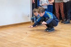 Dwóch chłopców kucając na drewnianej podłodze gra w kapsle. Na podłodze narysowany przy pomocy kredy tor dla kapsli. W tle nogi innych dzieci przyglądających się grze