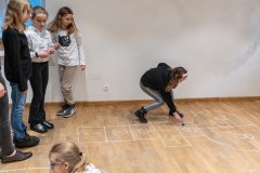 Dziewczynki grają w „chłopka”. Pola do gry narysowane kredą na terakocie. Jedna z nich podnosi z ziemi niebieską piłeczkę.