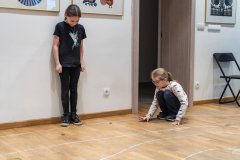 Dwie dziewczynki grają w kapsle. Jedna kuca, druga stoi obok. W tle przejście do innego pomieszczenia.
