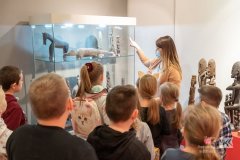 Pracownica Muzeum opowiada grupie dzieci o eksponatach znajdujących się w gablocie na wystawie
