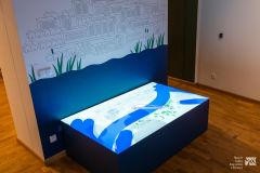 Film animowany o rzece Narew wyświetlony na poziomym ekranie wewnątrz sali wystawowej. Za poziomym ekranem, na ścianie, rysunek przedstawiający rzekę i zabudowania.