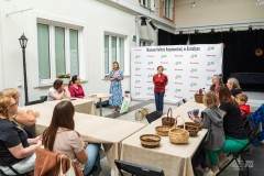 Przy dużym stole w kształcie litery "U" siedzą uczestnicy warsztatów. Przed nimi stoją dwie kobiety prowadzące spotkanie.