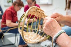 Dłonie wyplatające koszyk z wikliny. W tle dwie siedzące kobiety.