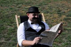 Mężczyzna z brodą i wązami, w meloniku, z muchą i monoklem leży na leżaku i czyta gazetę. W tle trawnik