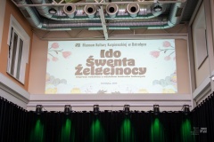 Na krytym dziedzińcu, na ścianie pod sufitem, wyświetlony banner z napisami: Muzeum Kultury Kurpiowskiej w Ostrołęce, Ido Śwenta Źelgejnocy, Impreza rodzinna z udziałem twórców ludowych, Ostrołęka, 2024