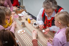 Przy stole, kobieta w stroju kurpiowskim pomaga dziewczynce w wykonaniu pisanki. Obok siedzi kilkoro innych dzieci malujących pisanki