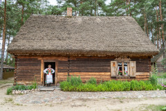 Kobieta w kurpiowskim stroju stoi w wejściu do kurpiowskiej chatki w skansenie.