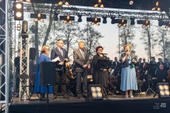 Trzy kobiety i dwóch mężczyzn na scenie zwracają się do widowni. W tle fragment orkiestry.