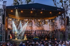 Chór z dyrygentem oraz soliści i solistki występujący na scenie. Z lewej strony na scenie siedzi kobieta w kurpiowskim stroju.