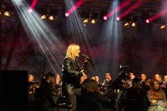 Wokalistka występująca na barwnie oświetlonej scenie. W tle fragment orkiestry
