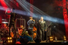 Dwóch wokalistów oraz dyrygent oraz fragment orkiestry na barwnie oświetlonej scenie.