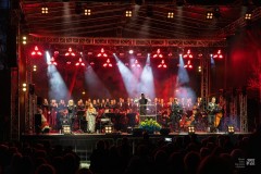 Wokaliści, orkiestra i chór z dyrygentem występujący na scenie. Z lewej strony na scenie siedzi kobieta w kurpiowskim stroju. Scena barwnie oświetlona