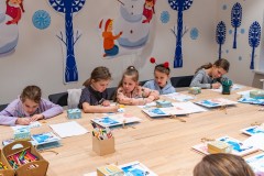 Pięcioro dzieci siedzących przy stole i rozwiązujących karty edukacyjne. W tle, na ścianie zimowa dekoracja z motywem wycinanki kurpiowskiej