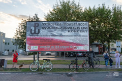 Billboard reklamujący koncert, w tle drzewa, fragment zabudowań. O bilboard oparte dwa rowery, w pobliżu stoi kilka osób, w tym jeden człowiek z rowerem.