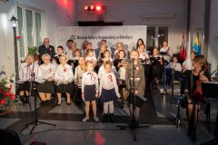 Grupa dzieci i dorosłych siedzi, dziewczynka przemawia do mikrofonu.