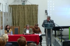 Mężczyzna stoi przy mównicy, obok dwie kobiety i mężczyzna siedzą przy stole nakrytym czerwonym obrusem.