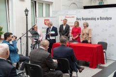 Prowadzący konferencję stoją za mównicą. Z lewej strony część widowni