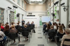 Na dziedzińcu w Muzeum Kultury Kurpiowskiej uczestnicy konferencji słuchają przemówienia. Na końcu sali stoi baner z logo Muzeum Kultury Kurpiowskiej w Ostrołęce.