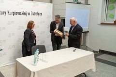 Kobieta stojąca za stołem i dwóch mężczyzn trzymają w rękach książki. W tle baner z logo Muzeum Kultury Kurpiowskiej.