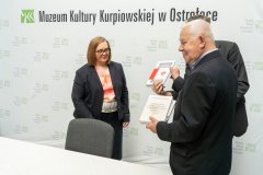 Pani dyrektor Muzeum Kultury Kurpiowskiej stoi z dwoma mężczyznami którzy trzymają w rękach książki . W tle baner z logo muzeum.