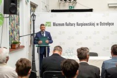 Stojący mężczyzna przemawia do kilku uczestników konferencji. Po lewej stronie są okna w kolorowych barwach natury a w tle baner z logo Muzeum Kultury Kurpiowskiej w Ostrołęce.