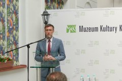 Mężczyzna stojący przed mównicą przemawia do zgromadzonych uczestników konferencji. W tle baner z logo Muzeum Kultury Kurpiowskiej w Ostrołęce. 