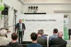 Kilku uczestników konferencji słucha przemówienia mężczyzny stojącego przy mównicy.