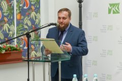 Mężczyzna stojący przy mównicy czyta prelekcję historyczną. W tle po lewej kolorowe okna a za mężczyzną baner z logo muzeum.  