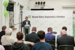 Stojący przy mównicy mężczyzna odczytuje referat historyczny.  W tle baner z logo Muzeum Kultury Kurpiowskiej w Ostrołęce.