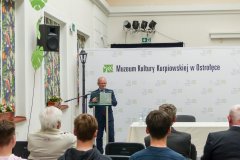 Stojący mężczyzna przy mównicy odczytuje prelekcje grupie uczestników konferencji historycznej. Po lewej stronie kolorowe okna a w tle baner z logo Muzeum Kultury Kurpiowskiej.
