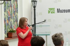 Kobieta w czerwonej sukience, stojąca bokiem i patrząca w ekran przemawia do uczestników konferencji. Za nią kolorowe okna a w tle baner z logo Muzeum Kultury Kurpiowskiej.