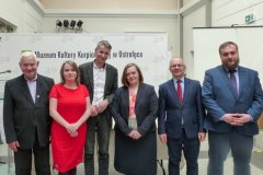 Dwie kobiety i czterech mężczyzn pozujących do zdjęcia stoi na dziedzińcu Muzeum Kultury Kurpiowskiej na  tle banera muzeum.                                         tle banera z logo Muzeum Kultury Kurpiowskiej.