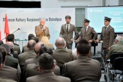 Mężczyzna stoi przy mikrofonie obok 3 mężczyźni przed widownią w tle napis Muzeum Kultury Kurpiowskiej