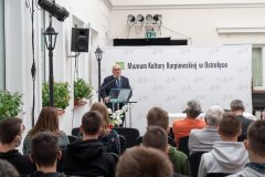 Uczestnicy konferencji słuchają przemówienia prelegenta stojącego przy mównicy z lewej strony sali. W tle ścianka z napisem Muzeum Kultury Kurpiowskiej w Ostrołęce oraz logotypami Muzeum.