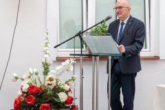 Prelegent stoi przy mównicy. Na ziemi, przy mównicy stoi biało-czerwony bukiet kwiatów.