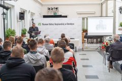 Uczestnicy konferencji słuchają przemówienia prelegenta stojącego przy mównicy z lewej strony sali. W tle ścianka z napisem Muzeum Kultury Kurpiowskiej w Ostrołęce oraz logotypami Muzeum. Z prawej strony ekran z wyświetloną prezentacją.