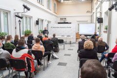 Uczestnicy konferencji słuchają przemówienia prelegenta stojącego przy mównicy z lewej strony sali. W tle ścianka z napisem Muzeum Kultury Kurpiowskiej w Ostrołęce oraz logotypami Muzeum. Z prawej strony ekran z wyświetloną prezentacją.
