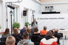 Uczestnicy konferencji słuchają przemówienia prelegenta stojącego przy mównicy z lewej strony sali. W tle ścianka z napisem Muzeum Kultury Kurpiowskiej w Ostrołęce oraz logotypami Muzeum.