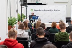 Uczestnicy konferencji słuchają przemówienia prelegenta stojącego przy mównicy z lewej strony sali. W tle ścianka z napisem Muzeum Kultury Kurpiowskiej w Ostrołęce oraz logotypami Muzeum.