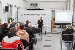 Uczestnicy konferencji słuchają przemówienia prelegentki stojącej przed widownią. W tle ścianka z napisem Muzeum Kultury Kurpiowskiej w Ostrołęce oraz logotypami Muzeum. Z prawej strony ekran z wyświetloną prezentacją.